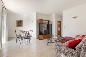 Uma sala de estar com sofá, mesa de centro de vidro, rack para TV e um canto de jantar com cadeiras e mesa.