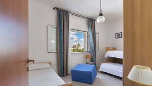 Een kamer met twee bedden, een blauwe poef, een tafel met een lamp en een raam met gordijnen.