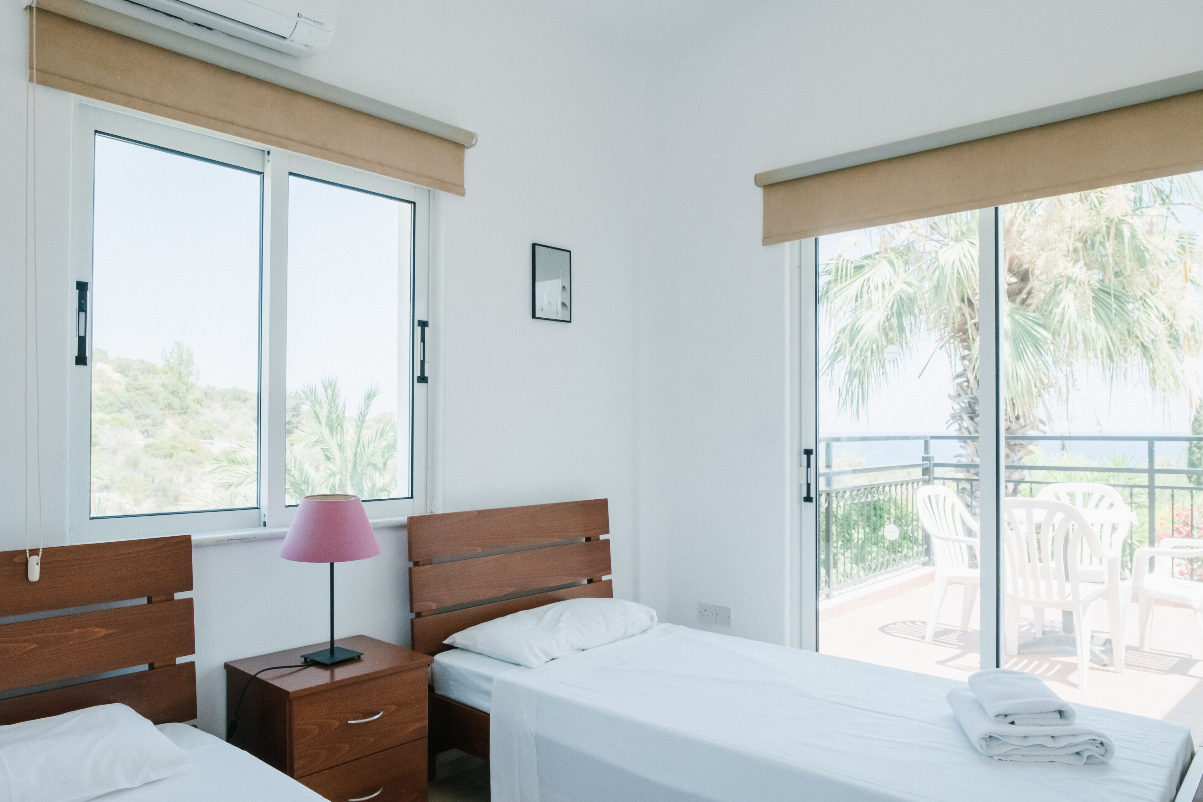 A bedroom with two beds, a nightstand, a lamp, and a balcony with chairs.