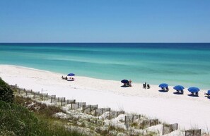 A beach with white sand and clear turquoise water. There are several blue umbrellas and beach chairs lined up along the shore.