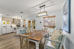 A dining area with a wooden table and chairs, a kitchen in the background, and a large window with blinds.