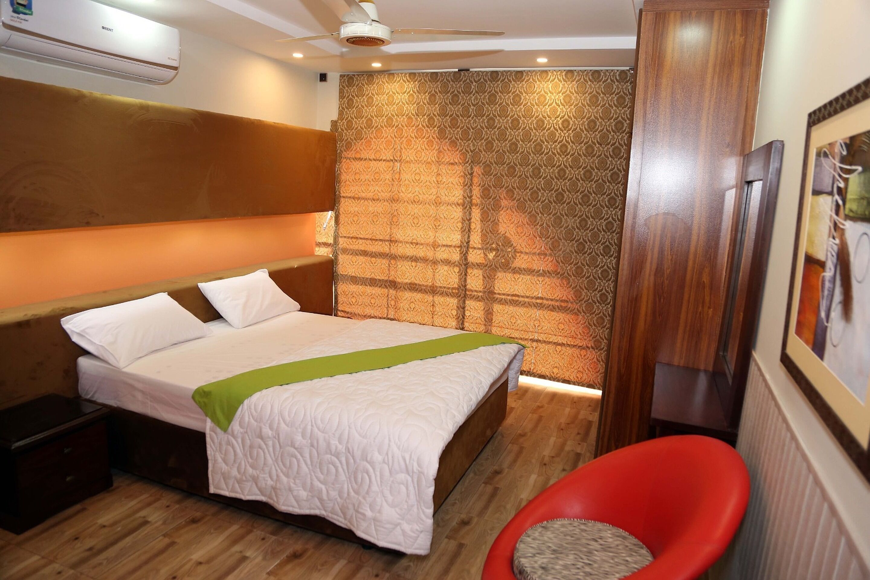A hotel room with a bed, a red chair, a wooden headboard, a patterned wall, and a ceiling fan.