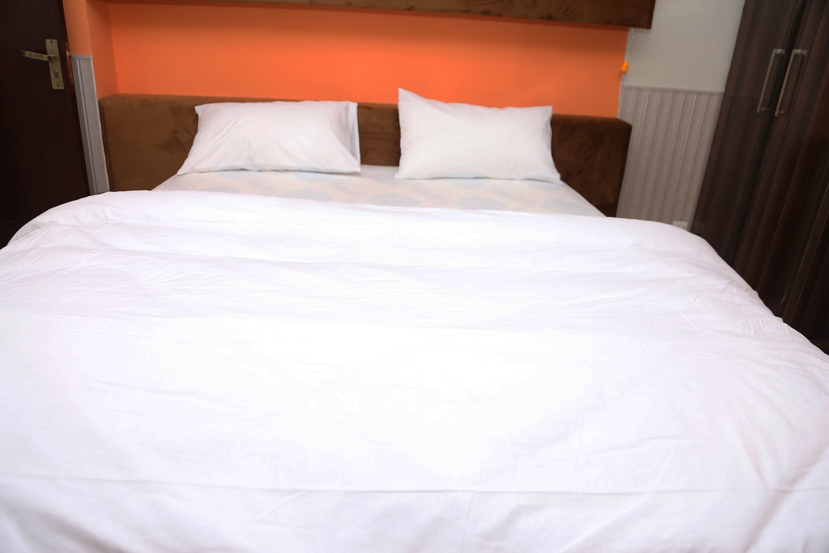 Un lit bien fait, avec du linge de lit blanc et deux oreillers, une tête de lit en bois et un mur d’accent de couleur marron.