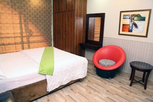 Una habitación con una cama, una silla roja, una mesita, un espejo y un cuadro enmarcado en la pared.