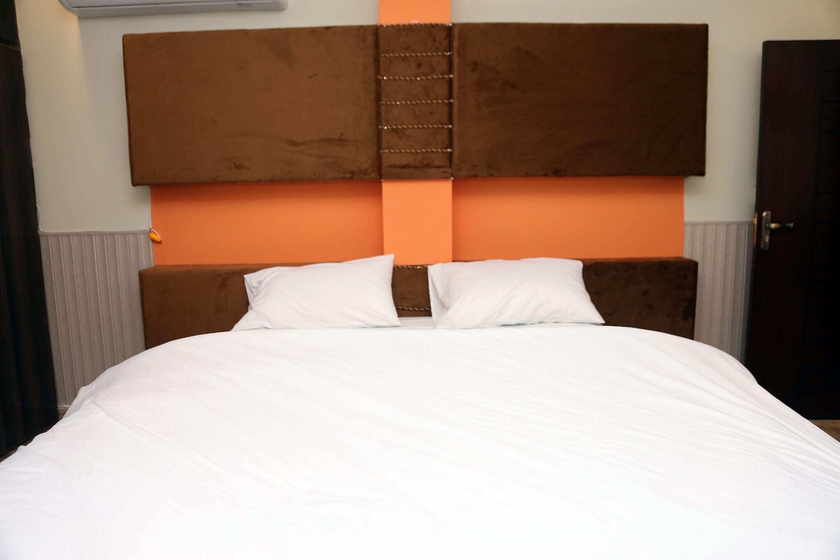 A hotel room with a large bed, a brown headboard, and an orange accent wall.