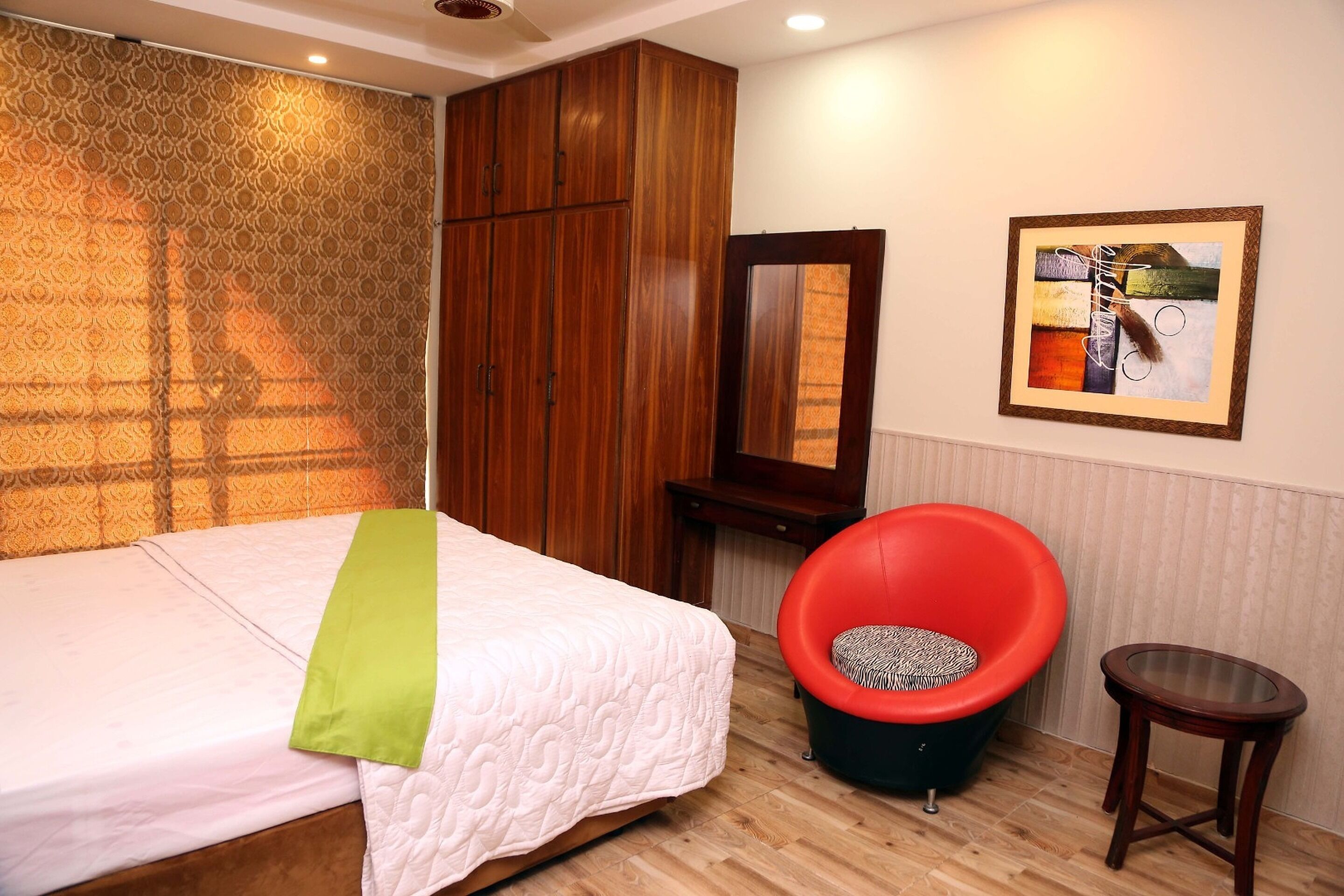 A bedroom with a bed, a red chair, a small table, a mirror, and a framed picture on the wall.