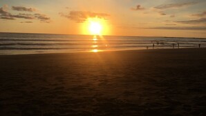 Een strand bij zonsondergang, met de zon laag aan de horizon, golven die rustig op het strand komen en een paar mensen die over het strand wandelen.