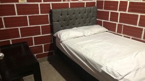 Una cama individual con cabecera capitonada frente a una pared de ladrillo, una mesita de noche de madera y una colcha blanca sencilla.