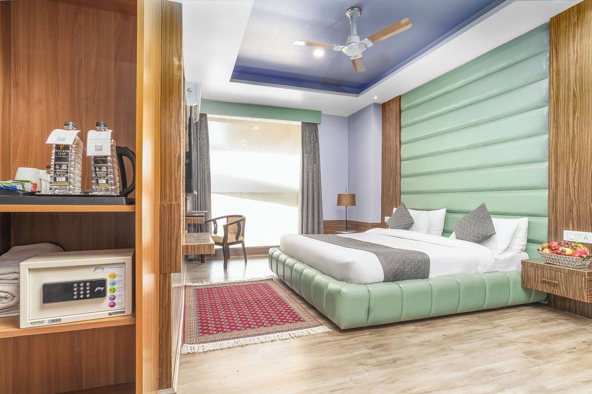 Habitación de hotel moderna con una cama tapizada de verde, un escritorio de madera con un microondas, un ventilador de techo y vistas a la ciudad a través de amplios ventanales.