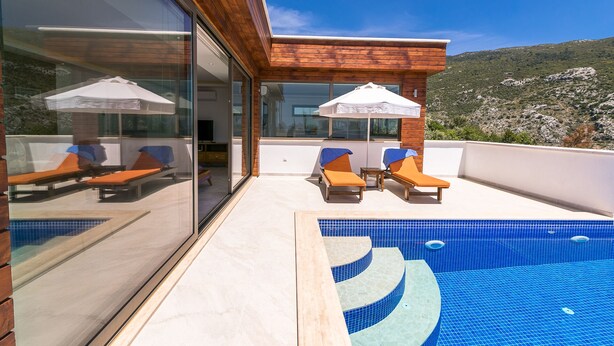 A modern outdoor pool area with lounge chairs and umbrellas, surrounded by a wooden deck and glass walls, overlooking a mountainous landscape.