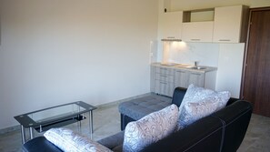Un espace de vie compact comprenant un canapé, une table basse en verre et, au fond, un coin cuisine.