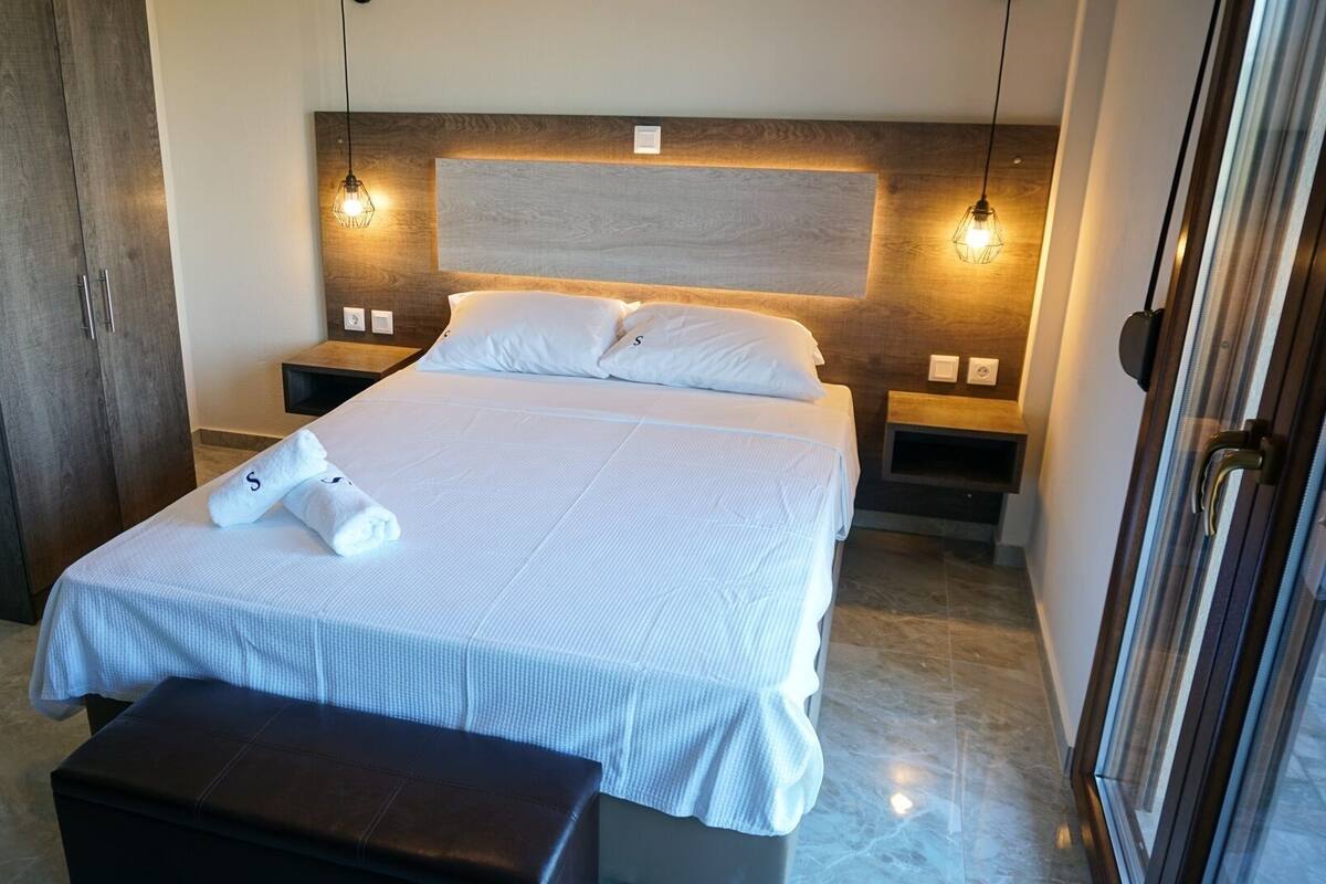 Ein ordentlich bezogenes Bett mit weißer Bettdecke und zwei Kissen, ein Holzkopfteil, Nachttische mit Lampen und eine Sitzbank am Fußende des Bettes.