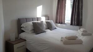 Cama bem arrumada com roupa de cama branca, almofadas cinzas e um par de toalhas dobradas.