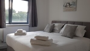 Uma cama bem arrumada com almofadas e toalhas dobradas, um radiador, uma janela com cortinas e um quadro na parede.