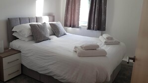 Cama arrumada com almofadas e toalhas dobradas, mesa de cabeceira com abajur e janela com cortinas.