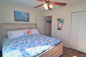 A bedroom with a bed, a ceiling fan, a door, and a wall with a palm tree decoration.