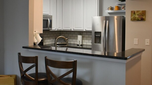 A kitchen with white cabinets, a stainless steel refrigerator, a microwave, and a sink. There are two wooden bar stools and a wooden table. - Special Pricing Uptown Condo Minutes from Bank of America Stadium (Charlotte)