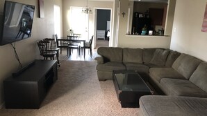 Una sala de estar con un sofá modular, una mesa de centro, un televisor sobre un mueble bajo y un comedor con sillas y una mesa.