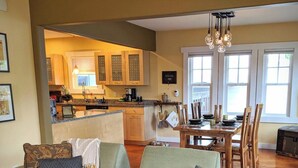 A cozy living room with a sofa, a dining table, and chairs. There is a kitchen in the background with cabinets and a window. - Dragonfly House-One Bedroom Craftsman in Trendy Kenton (Portland)