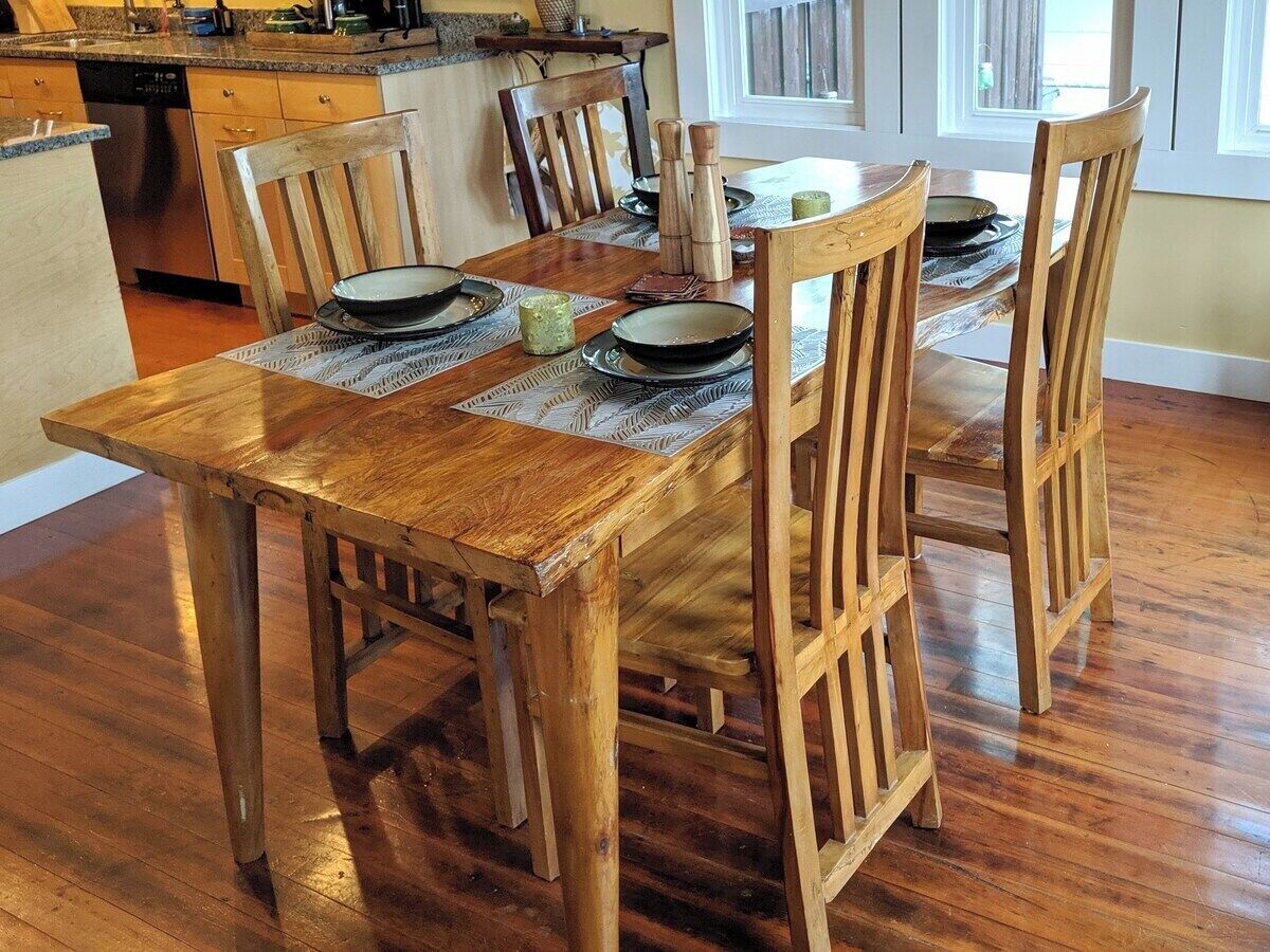 A wooden dining table with four chairs, a kitchen in the background, and a window with blinds.