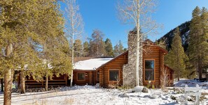Une cabane en rondins avec une cheminée en pierre, entourée d’un paysage enneigé et d’arbres.