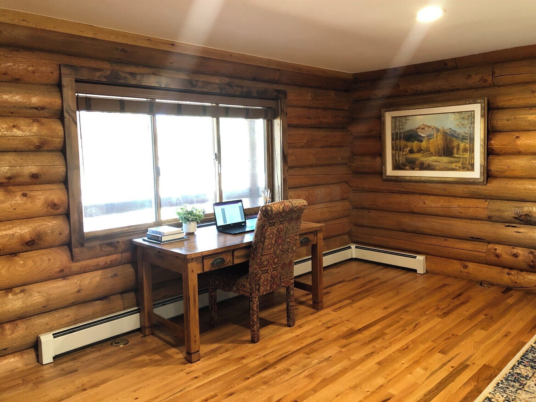 A log cabin room with a wooden desk, a patterned chair, a laptop, and a framed landscape painting on the wall.