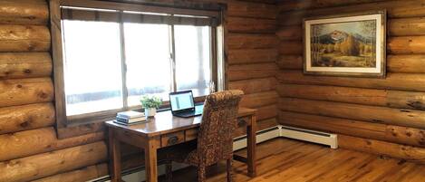 A log cabin room with a wooden desk, a patterned chair, a laptop, and a framed landscape painting on the wall.
