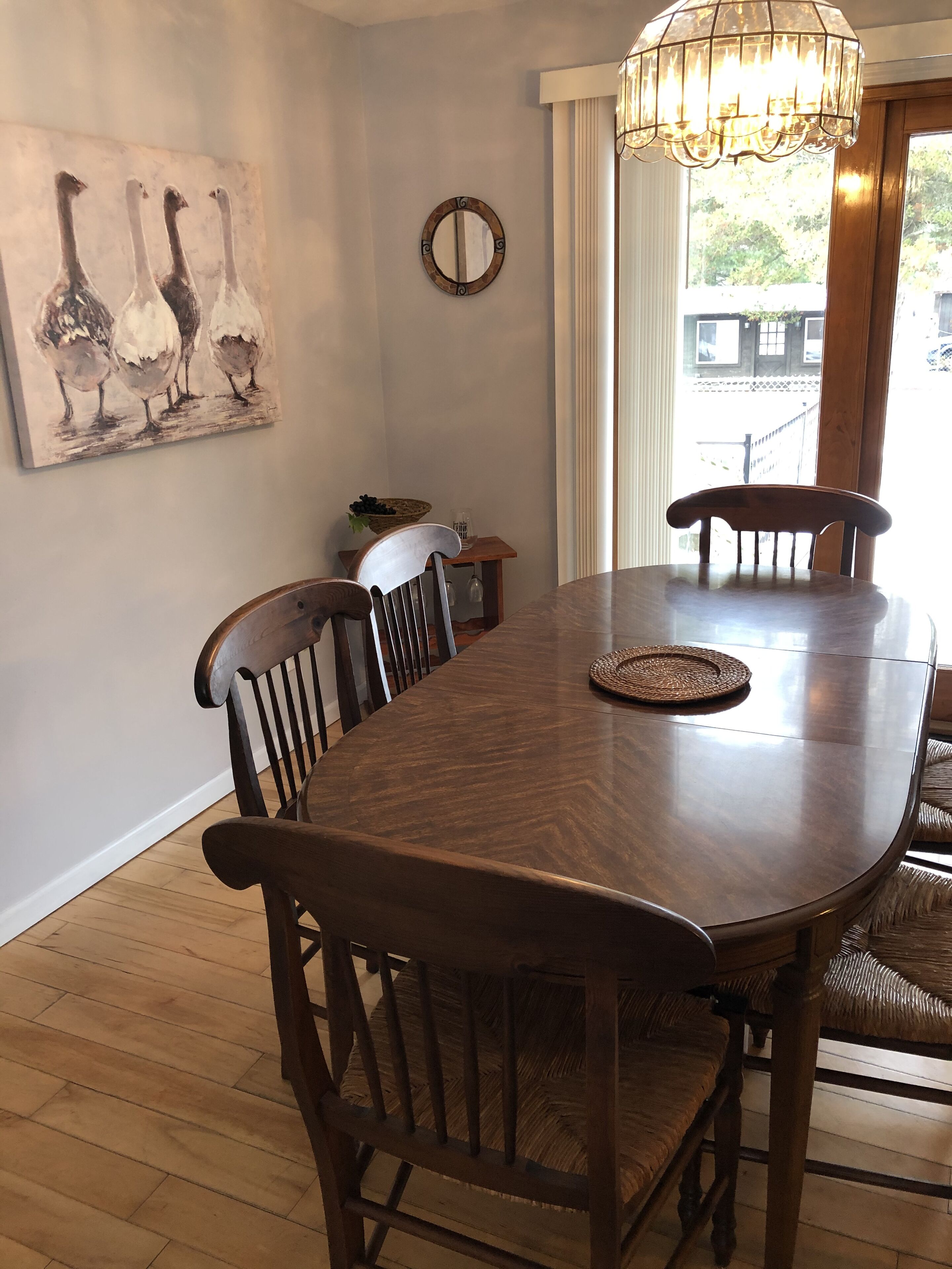 Un coin repas avec une table et des chaises en bois, un lustre, un tableau représentant des oies et une fenêtre avec des rideaux.