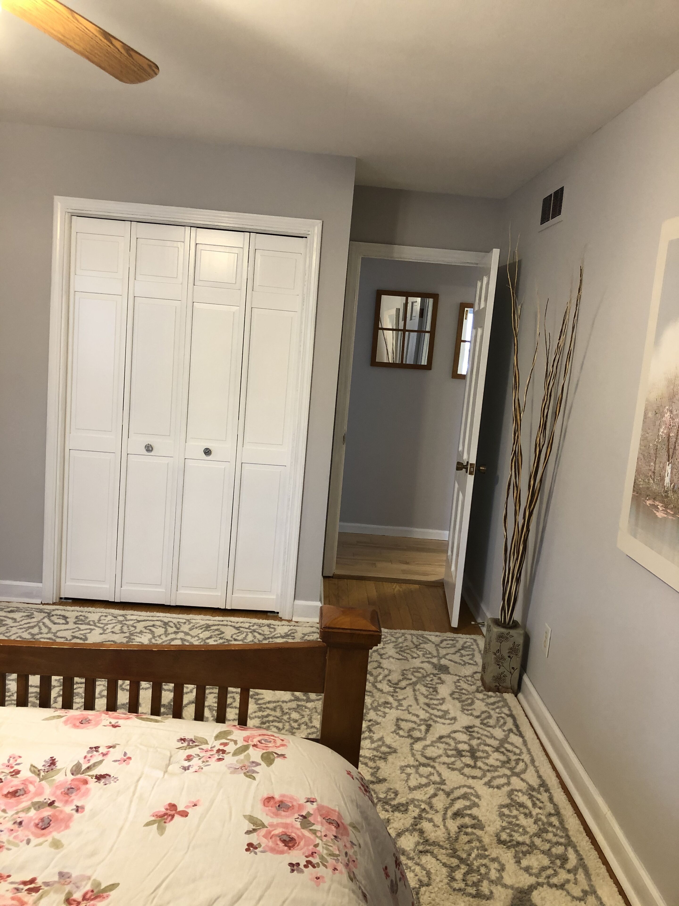 A bedroom with a bed, a nightstand, a white wardrobe, a mirror, and a vase with branches.