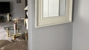 Une pièce avec un miroir encadré de blanc accroché au mur, un canapé beige, une table basse en verre ornée d’un vase de fleurs et un téléviseur en arrière-plan.