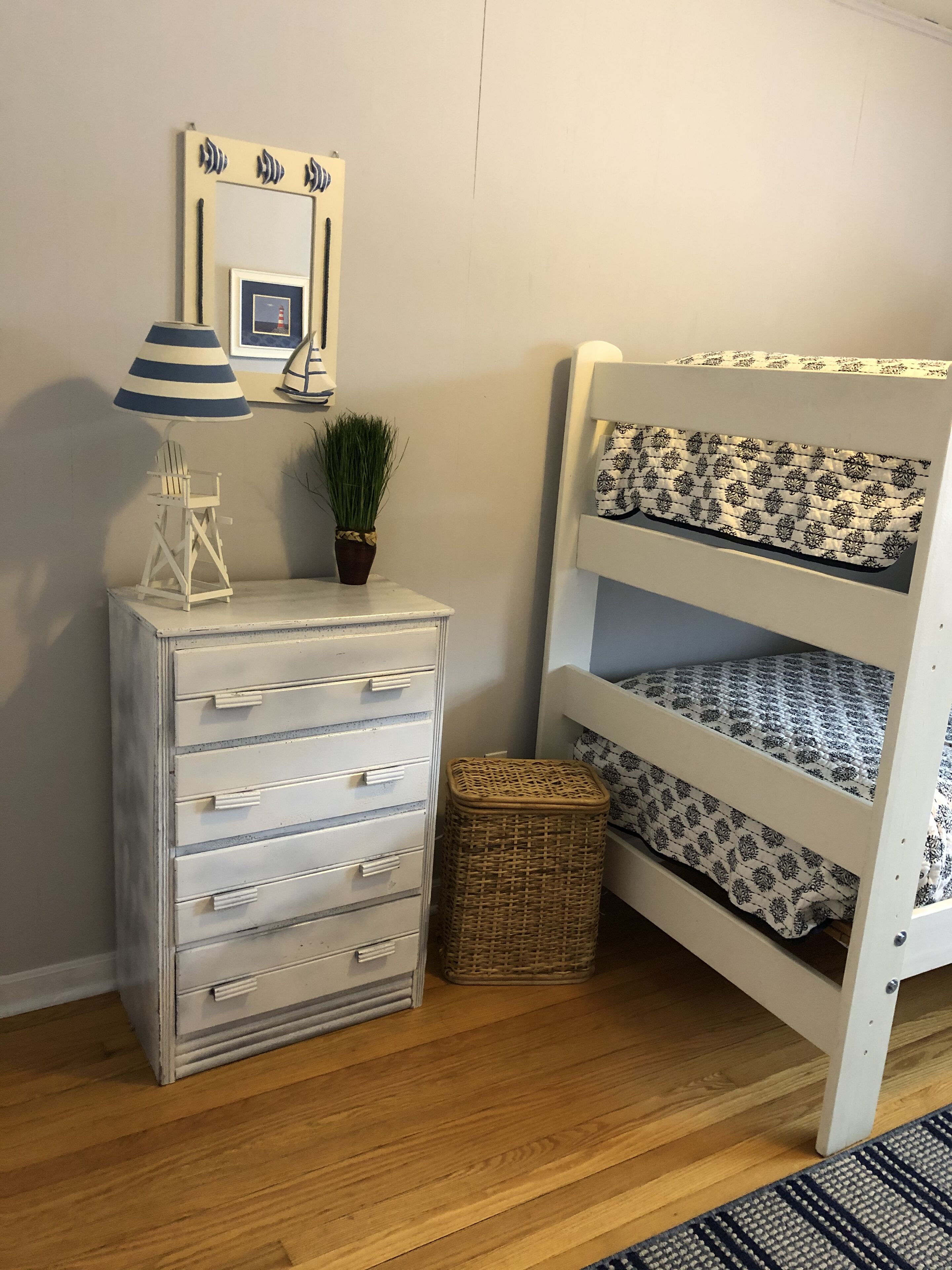 A bunk bed with patterned bedding, a dresser with drawers, a lamp, a potted plant, and a framed picture on the wall.