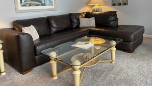 Un salon avec un canapé d’angle en cuir noir, une table basse en verre, une lampe et un tableau encadré accroché au mur.