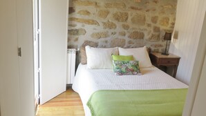 Een slaapkamer met een stenen muur, een bed met kussens, een nachtkastje met een lamp en een houten vloer.