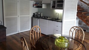 Een compacte keuken met witte kastjes, een magnetron en een wastafel. Twee rieten stoelen staan rond een glazen tafel met een groen voorwerp erop.