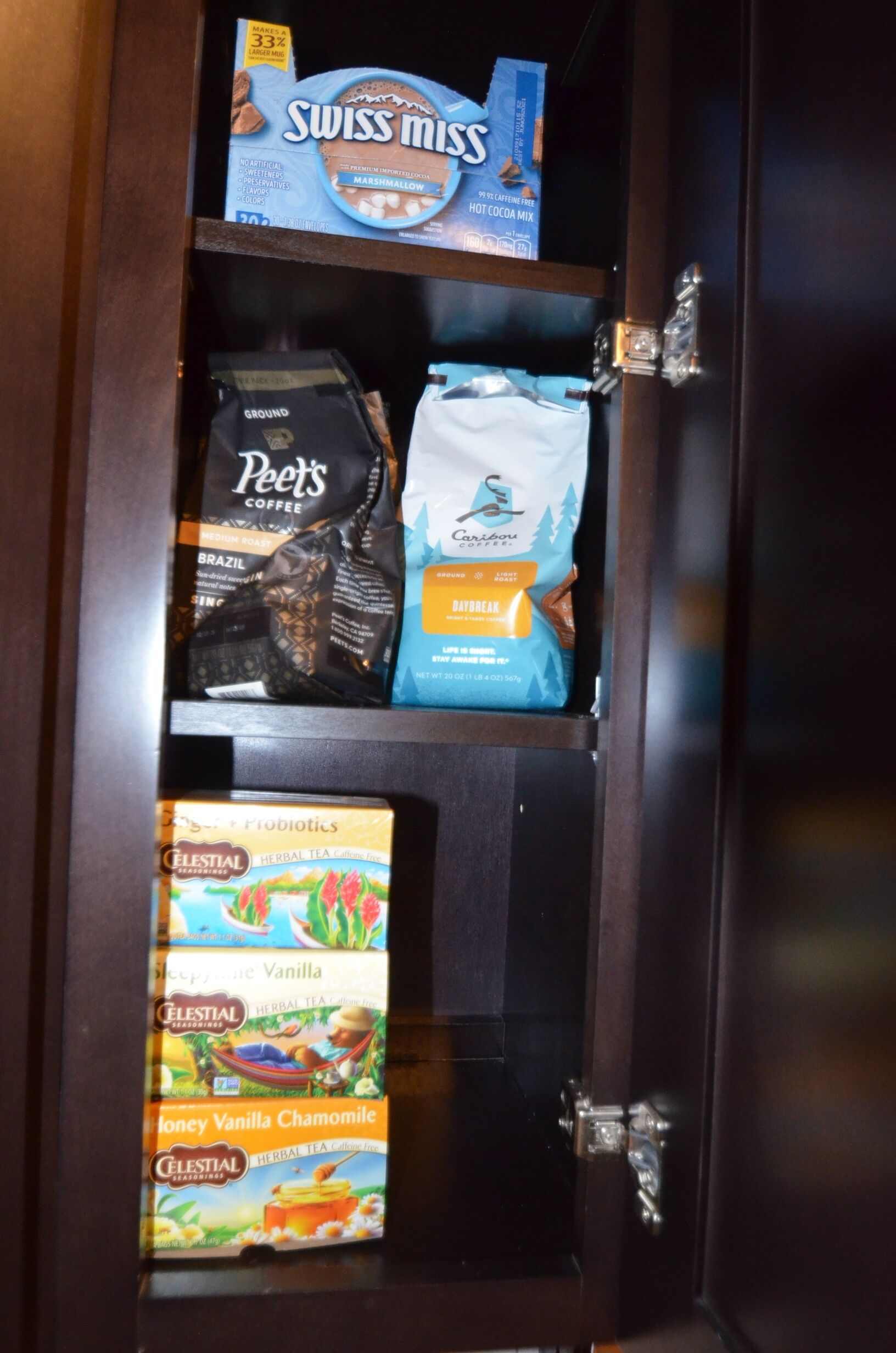 Un mueble con estantes que contiene varios productos alimenticios, incluyendo mezcla de malvaviscos Swiss Miss, café Peet's, café Caribou y tés herbales Celestial.