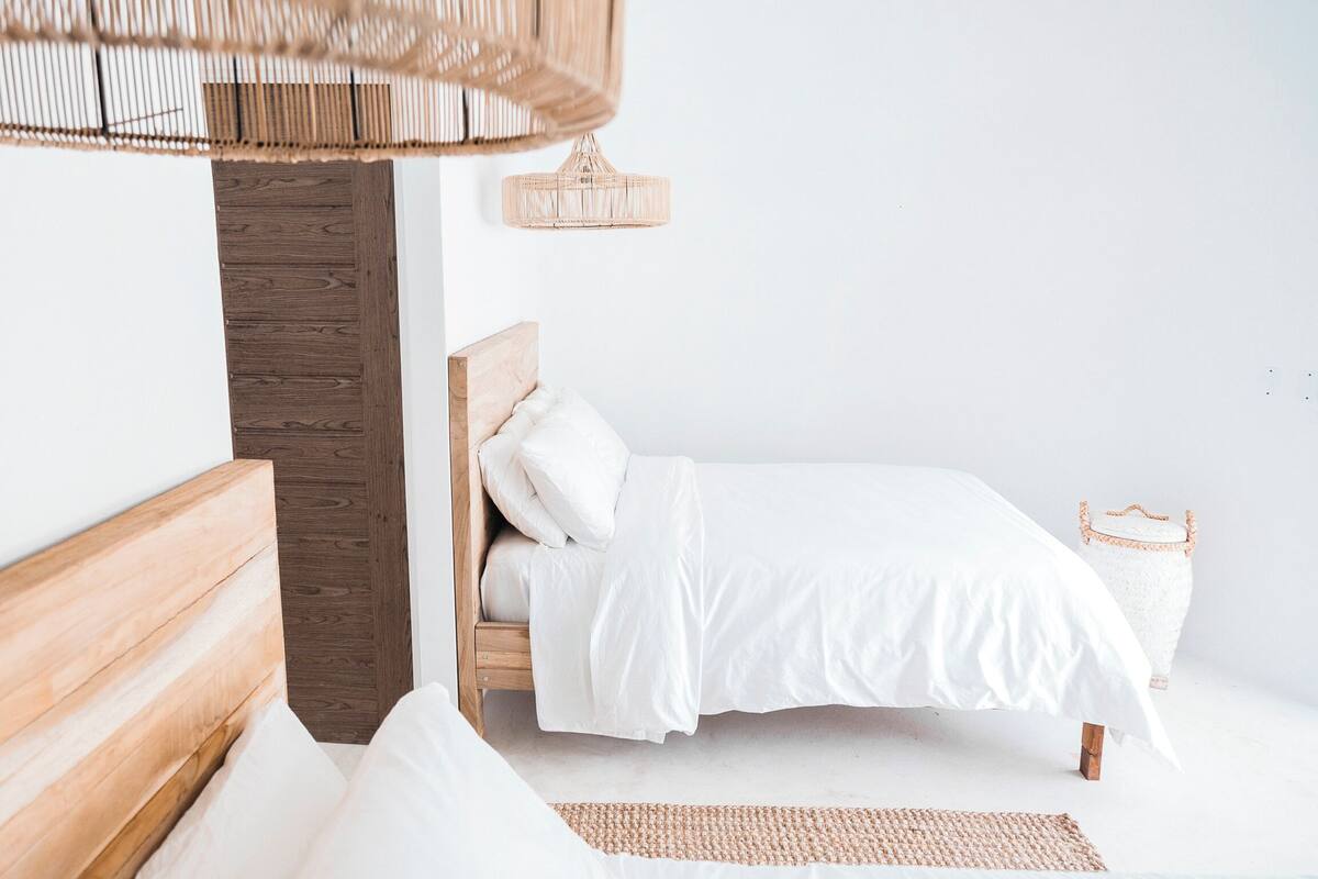 Quarto minimalista com cabeceira de madeira, cama com roupa de cama branca, tapete tecido e abajur de vime.