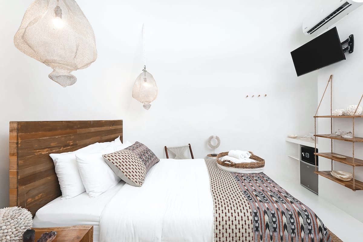 Um quarto moderno com cabeceira de madeira, uma cama grande com roupa de cama estampada, um lustre pendente, uma TV fixada na parede e um pequeno aparador.