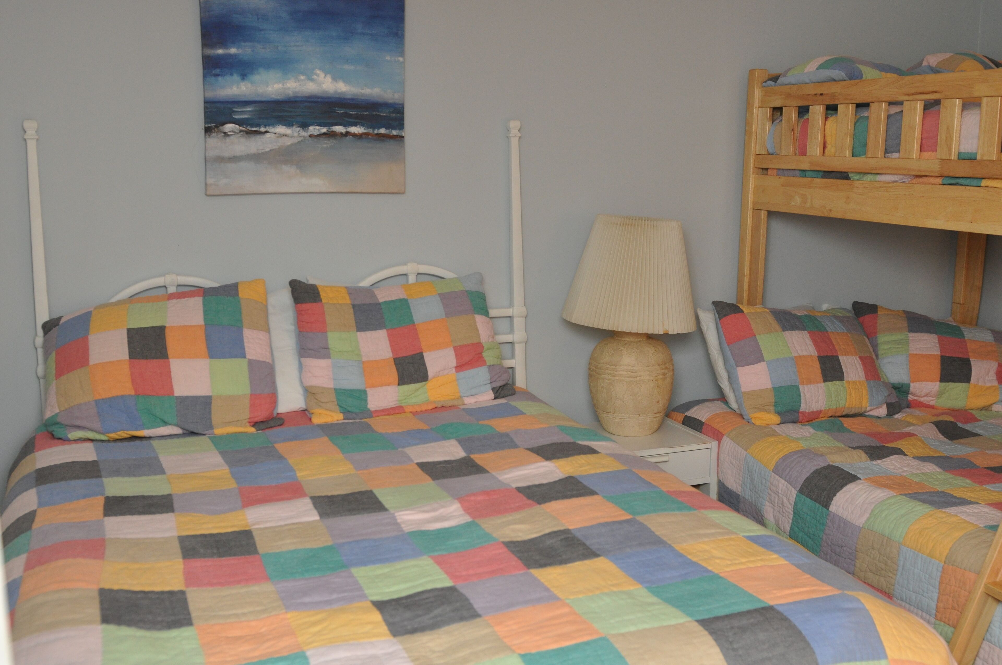 Une chambre avec deux lits, chacun agrémenté d’une literie à carreaux colorés, un lit superposé en bois, une lampe et un tableau représentant une scène de plage accroché au mur.