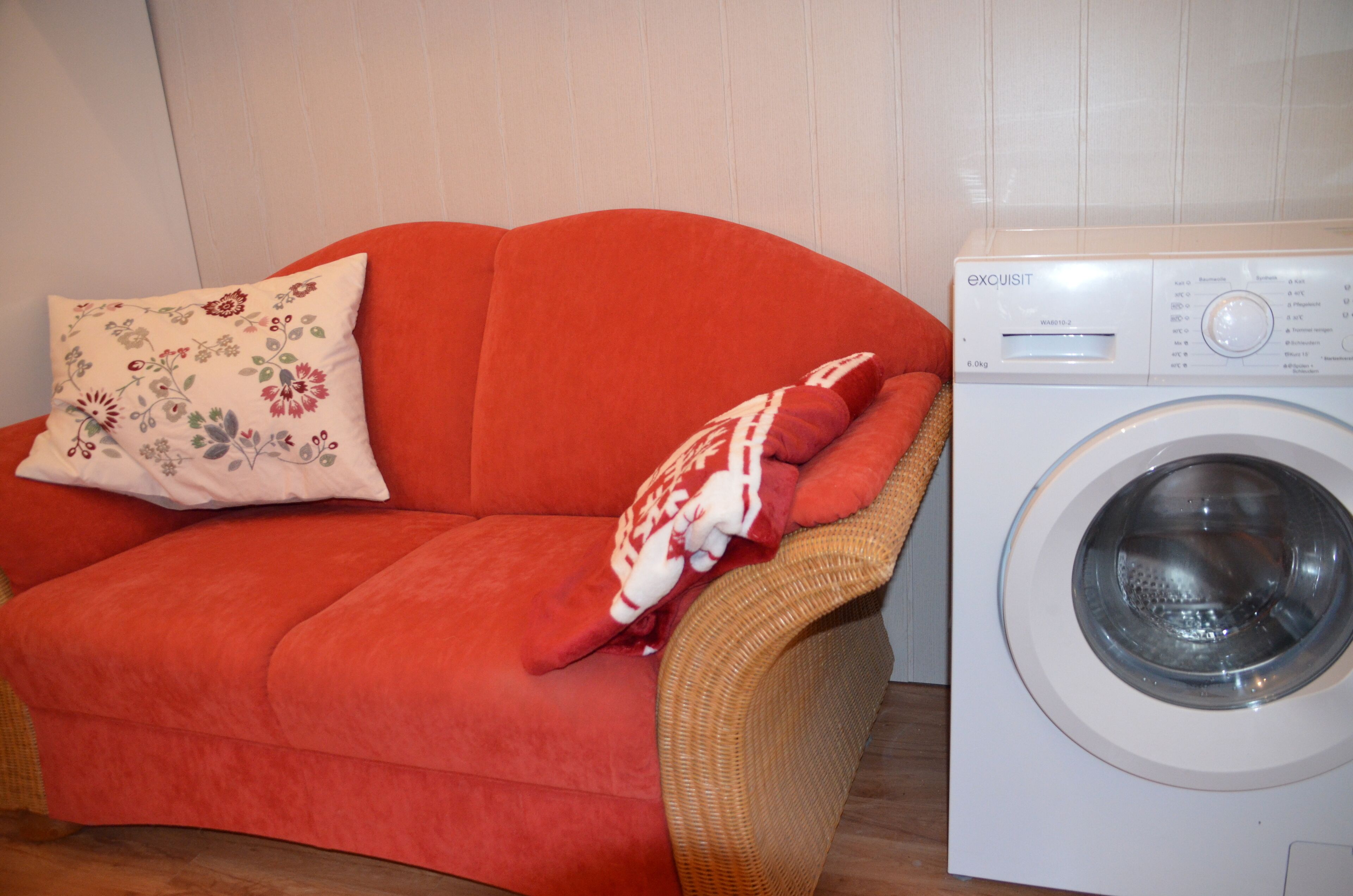 Un canapé rouge, orné d’un coussin à motifs floraux et recouvert d’une couverture, se trouve à côté d’un lave-linge blanc.