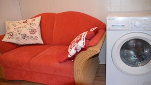 Un canapé rouge, orné d’un coussin à motifs floraux et recouvert d’une couverture, se trouve à côté d’un lave-linge blanc.