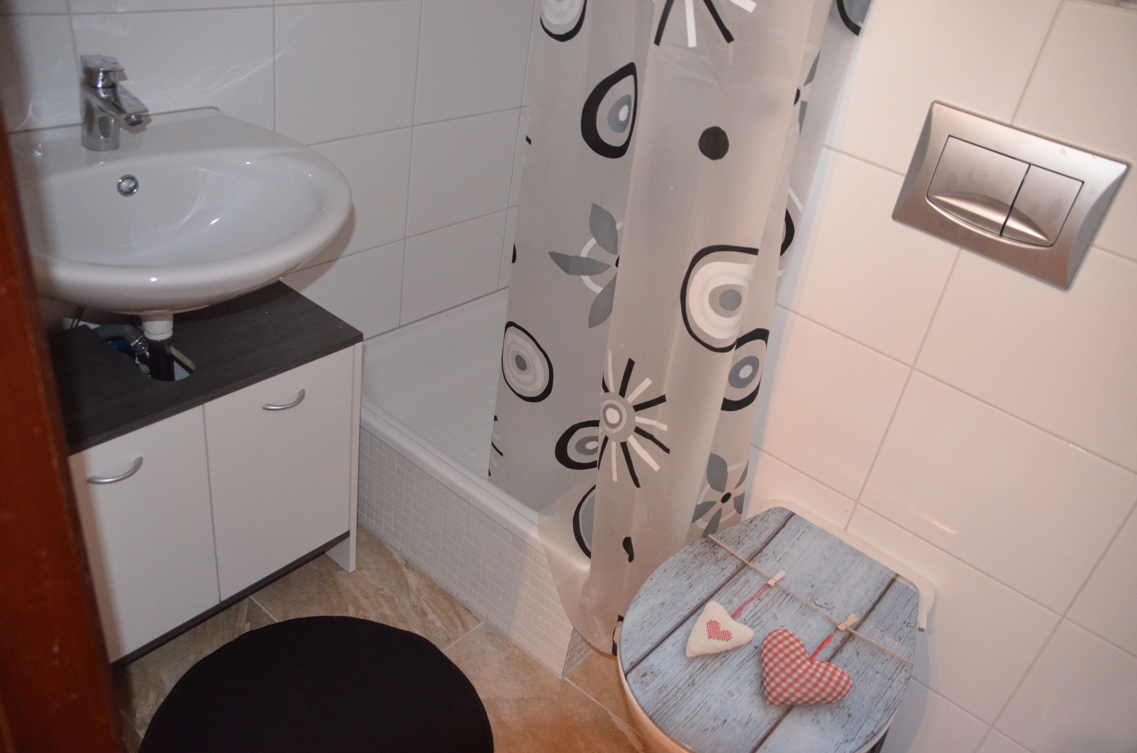 Um banheiro com pia branca, cortina de chuveiro com padrão floral, um banquinho de madeira pequeno com decoração de coração e um interruptor de luz fixado na parede.
