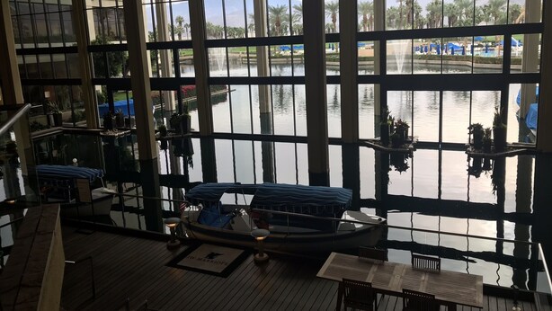A view from a high floor of a building with large windows overlooking a body of water, a dock with a boat, and a seating area with a table and chairs. - Beautiful Marriott Timeshare Unit located adjacent to JW Marriott (Palm Desert)