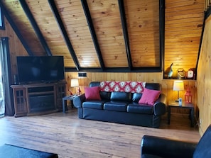 Un soggiorno accogliente con soffitto in legno, un divano in pelle nera con cuscini rossi e bianchi, una televisione su un mobile in legno, e un tavolino con una lampada e un lanterne rosso.