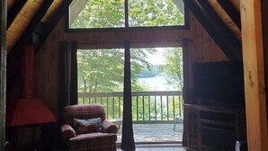 Une pièce mansardée chaleureuse avec une grande fenêtre, des poutres apparentes, un ventilateur de plafond, un fauteuil à carreaux, une lampe rouge et une cheminée.