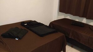 Two single beds with brown covers, a small table with a folded black jacket, and a window with brown curtains. - Ed. HSS (Ouro Preto)
