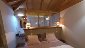 Una camera da letto con soffitto in legno, un ampio specchio, due abat-jour, un letto con lenzuola bianche e un comodino.