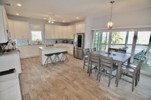 A modern kitchen with white cabinets, a central island with stools, a dining table with chairs, and a large window with a view of palm trees. - New luxury pool home, 2 min to beach, 360 water views, club available (North Captiva)