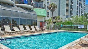 A swimming pool with lounge chairs, a fenced area, and a building with balconies in the background. - Gorgeous Oceanfront Condo With Direct Ocean View (Myrtle Beach)