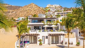 A beachfront hotel with a pool area, outdoor seating, and a view of the surrounding hillside. - Thanksgiving Promo!!  Your Exclusive Cabo Beachfront Escape (Cabo San Lucas)