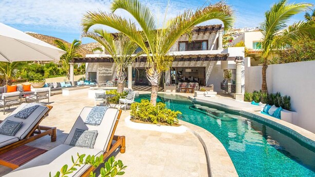 A pool area with lounge chairs, palm trees, and a building with a tiled roof. - Thanksgiving Promo!!  Your Exclusive Cabo Beachfront Escape (Cabo San Lucas)